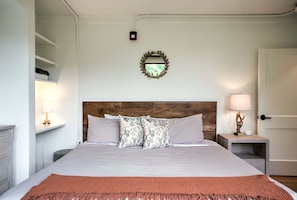 A neatly arranged bedroom with a bed, bedside tables, a nightstand, a wall-mounted light fixture, and a door. - Sabel #303 | Downtown Asheville king suite with kitchenette, Wi-Fi, parking. (Asheville)