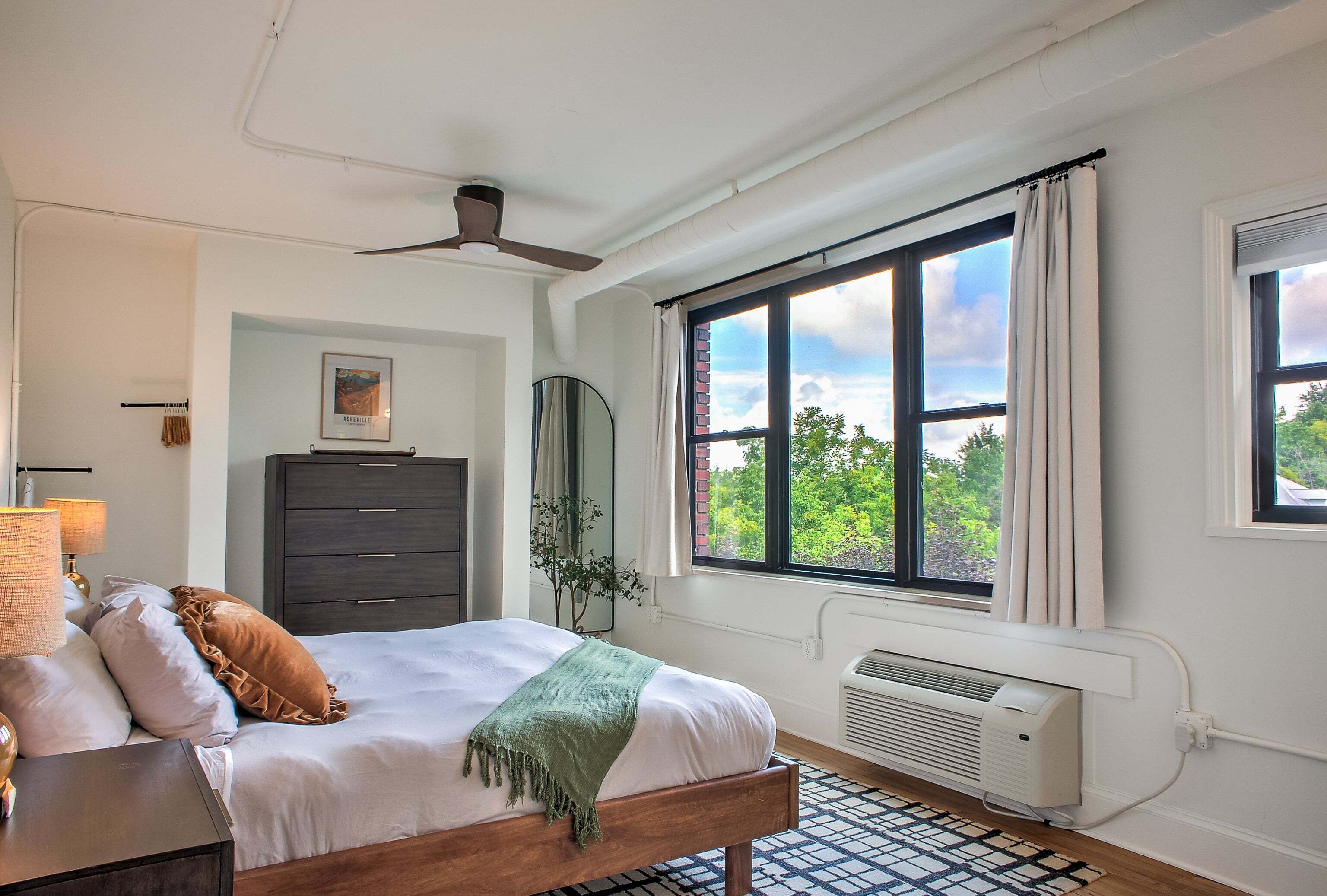 A bedroom with a bed, a dresser, a mirror, a ceiling fan, and a window with a view of trees.