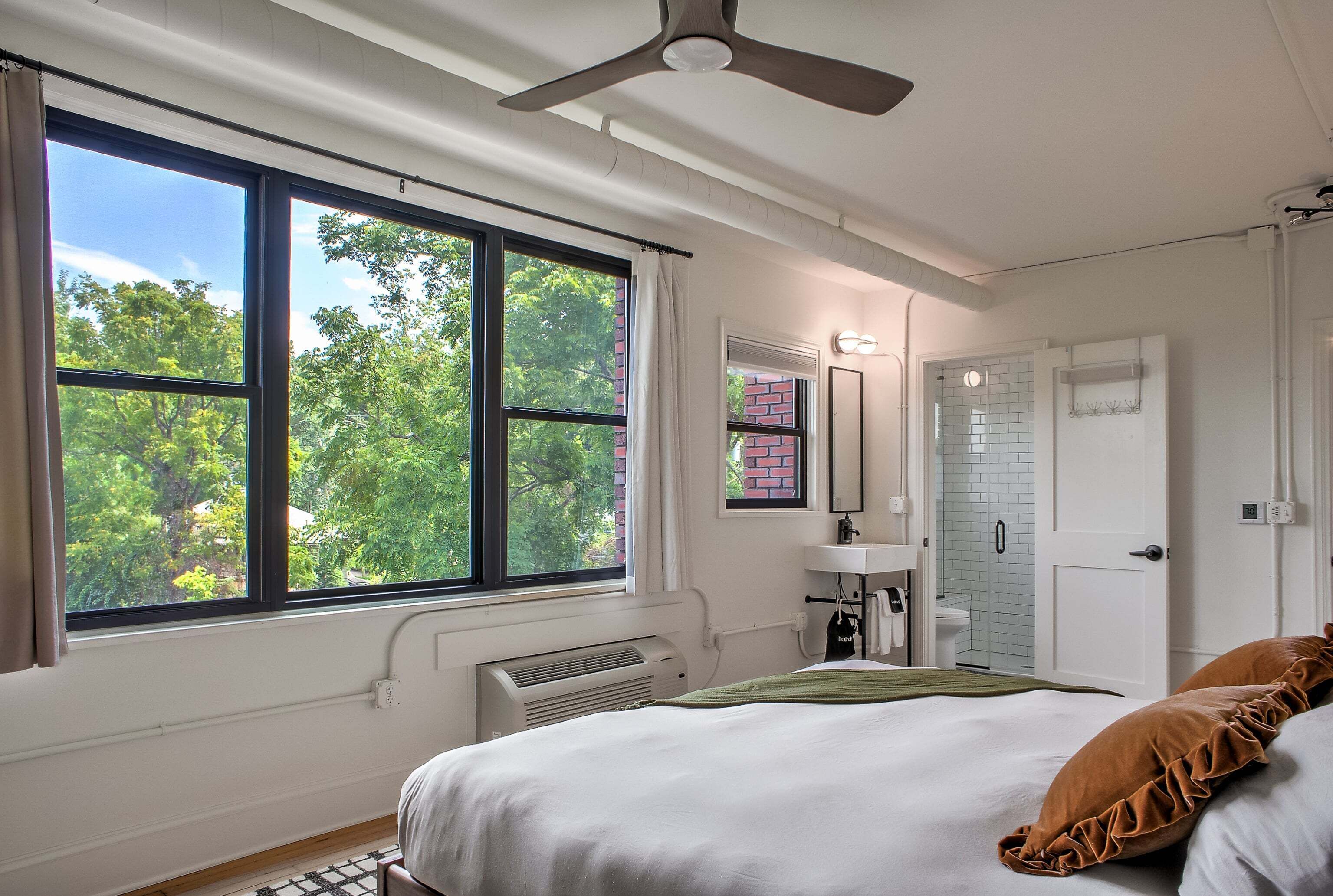 A bedroom with a large window, a ceiling fan, a radiator, a bed with pillows, a bathroom with a sink and mirror, and a door.