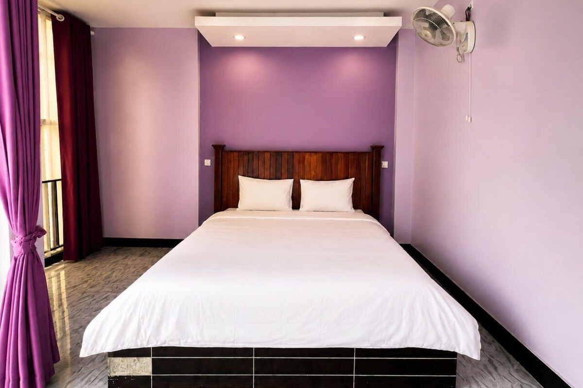 Habitació d'hotel amb un llit gran, un capçal de llit, un ventilador fixat a la paret i una finestra amb cortines.