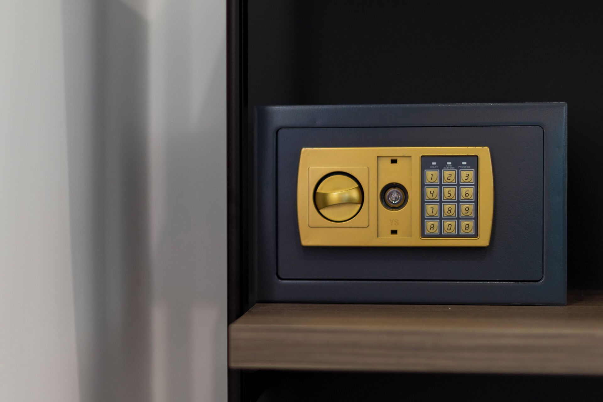 A safe with a combination dial and keypad, placed on a wooden shelf against a dark wall.