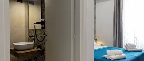 A bathroom with a sink, mirror, and hairdryer, and a bedroom with a bed, window, and curtains.