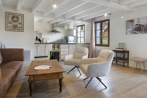 A spacious living area with a wooden coffee table, a brown leather sofa, and a beige armchair. A kitchen with white cabinets and a dining area with a wooden table and chairs is visible in the background.