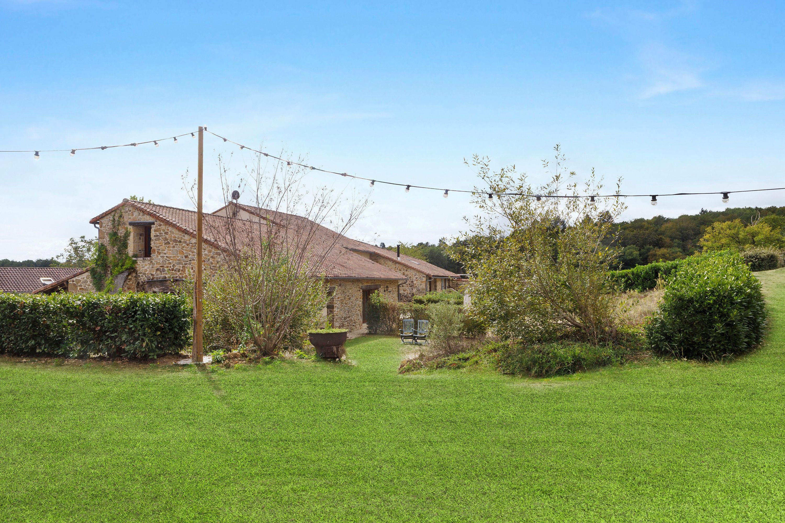 A stone house with a tiled roof, surrounded by a well-maintained lawn and a variety of shrubs and trees.