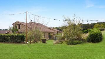 A stone house with a tiled roof, surrounded by a well-maintained lawn and a variety of shrubs and trees.