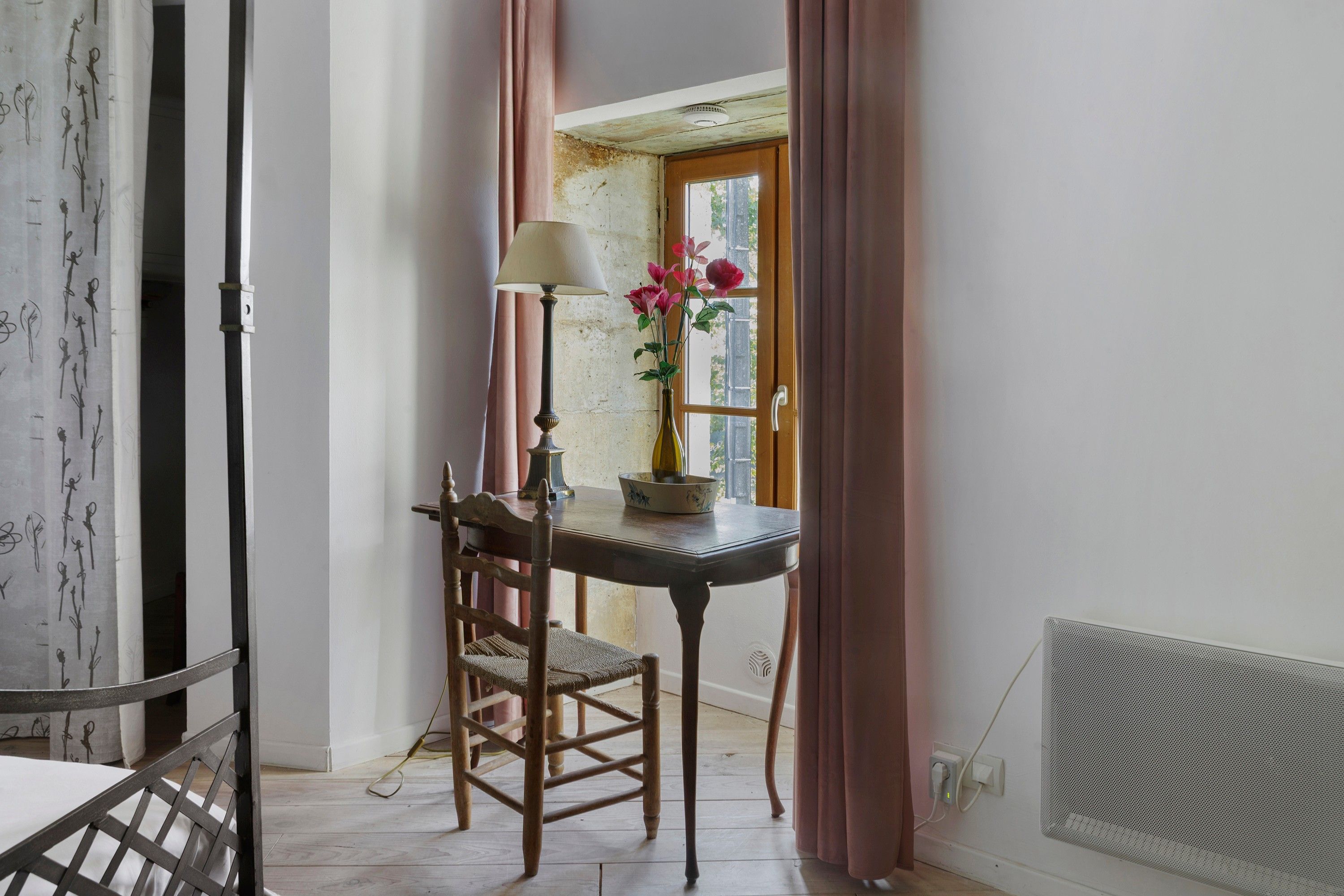 Une pièce avec une chaise en bois, une table avec une lampe, un vase rempli de fleurs et une fenêtre avec des rideaux.