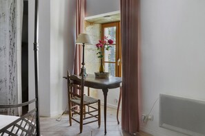 Una habitación con una silla de madera, una mesa con una lámpara, un florero con flores y una ventana con cortinas.