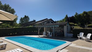 A swimming pool with lounge chairs, a covered patio, and a building with a tiled roof.