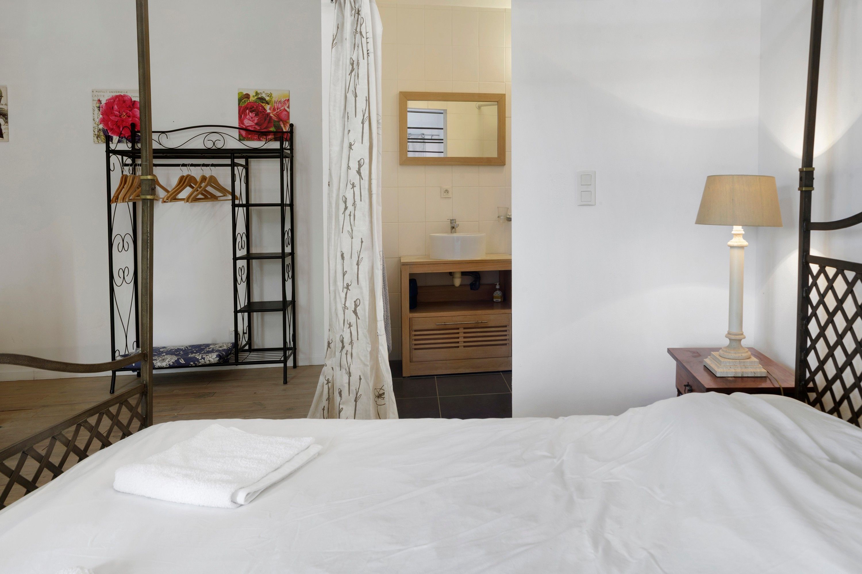 Une chambre à coucher comprenant un lit, une table de chevet avec une lampe, une commode en bois, un miroir et un portant à vêtements avec des cintres.