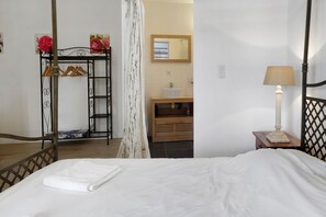 Un dormitorio con cama, mesita de noche con lámpara, cómoda de madera, espejo y perchero con perchas.