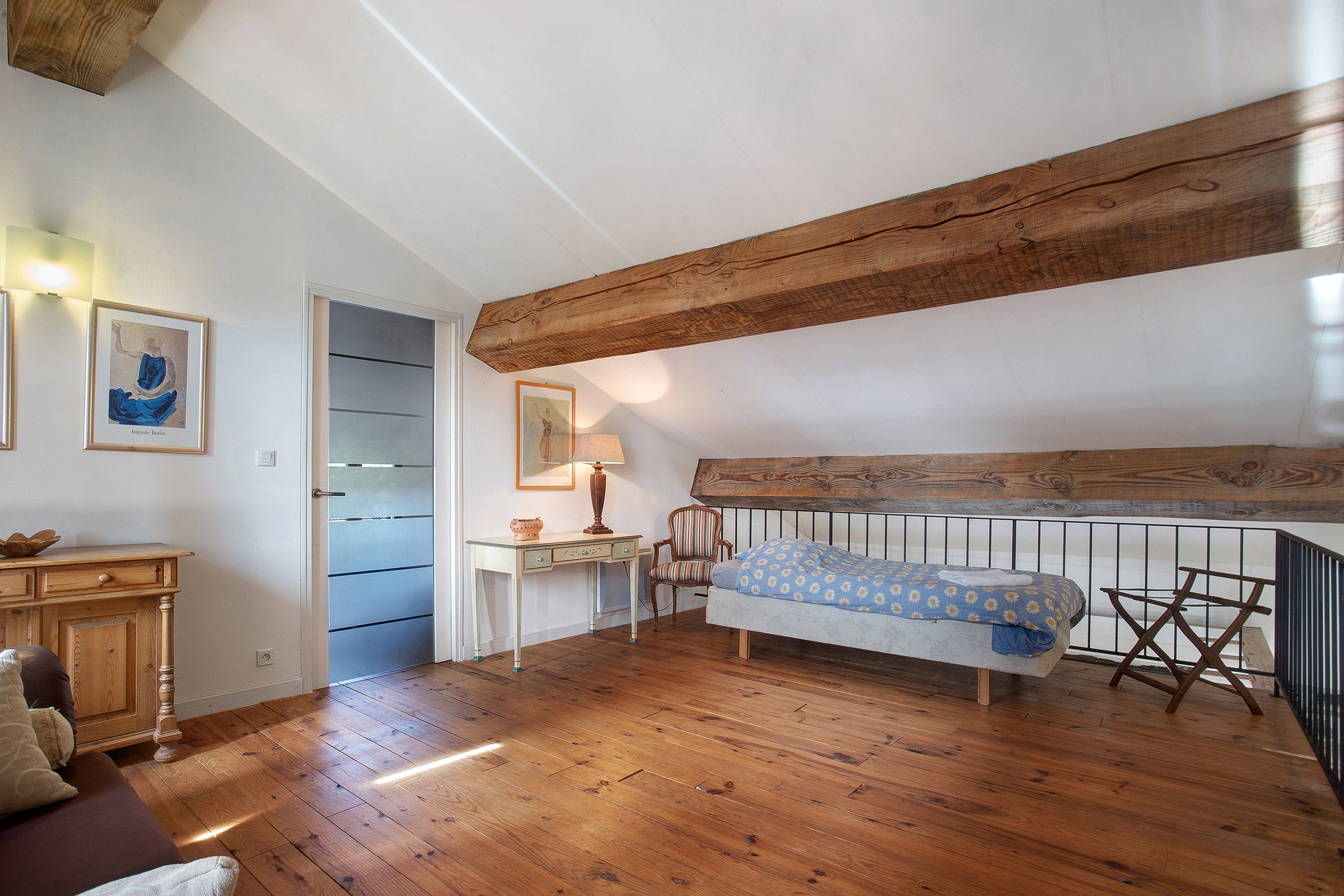 Een slaapkamer met een schuine kap, houten balken, een bed met een beddegoed met een patroon, een bureau met een lamp en een stoel.