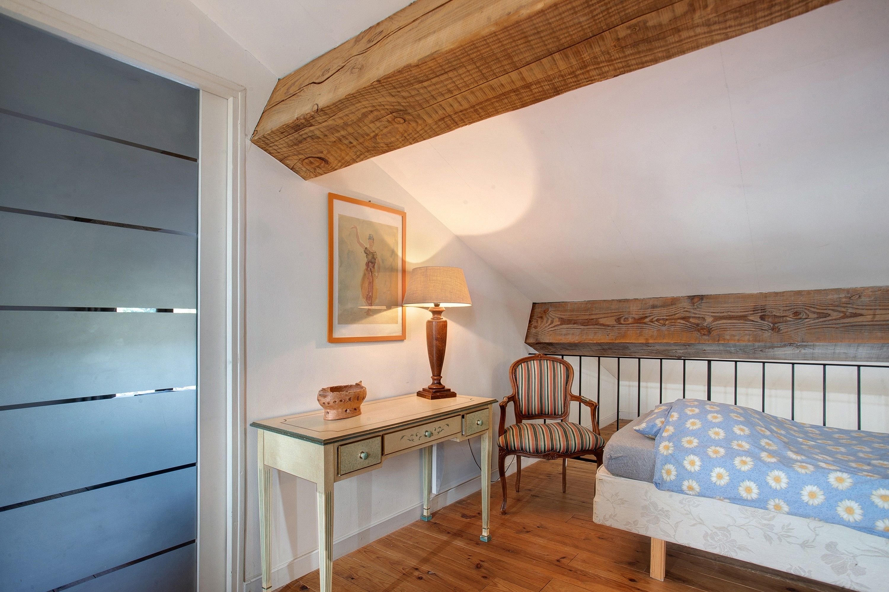Ein Schlafzimmer mit Holzboden, einem Bett mit blumigem Überwurf, einem Sessel, einem Schreibtisch mit Lampe und einem gerahmten Bild an der Wand.