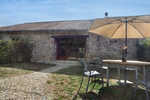 A stone building with a tiled roof, a wooden door, and a gravel driveway. There is a table with chairs and a large umbrella.