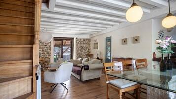 Ampla sala de estar com escada de madeira, parede de pedra e área de jantar com mesa de vidro.