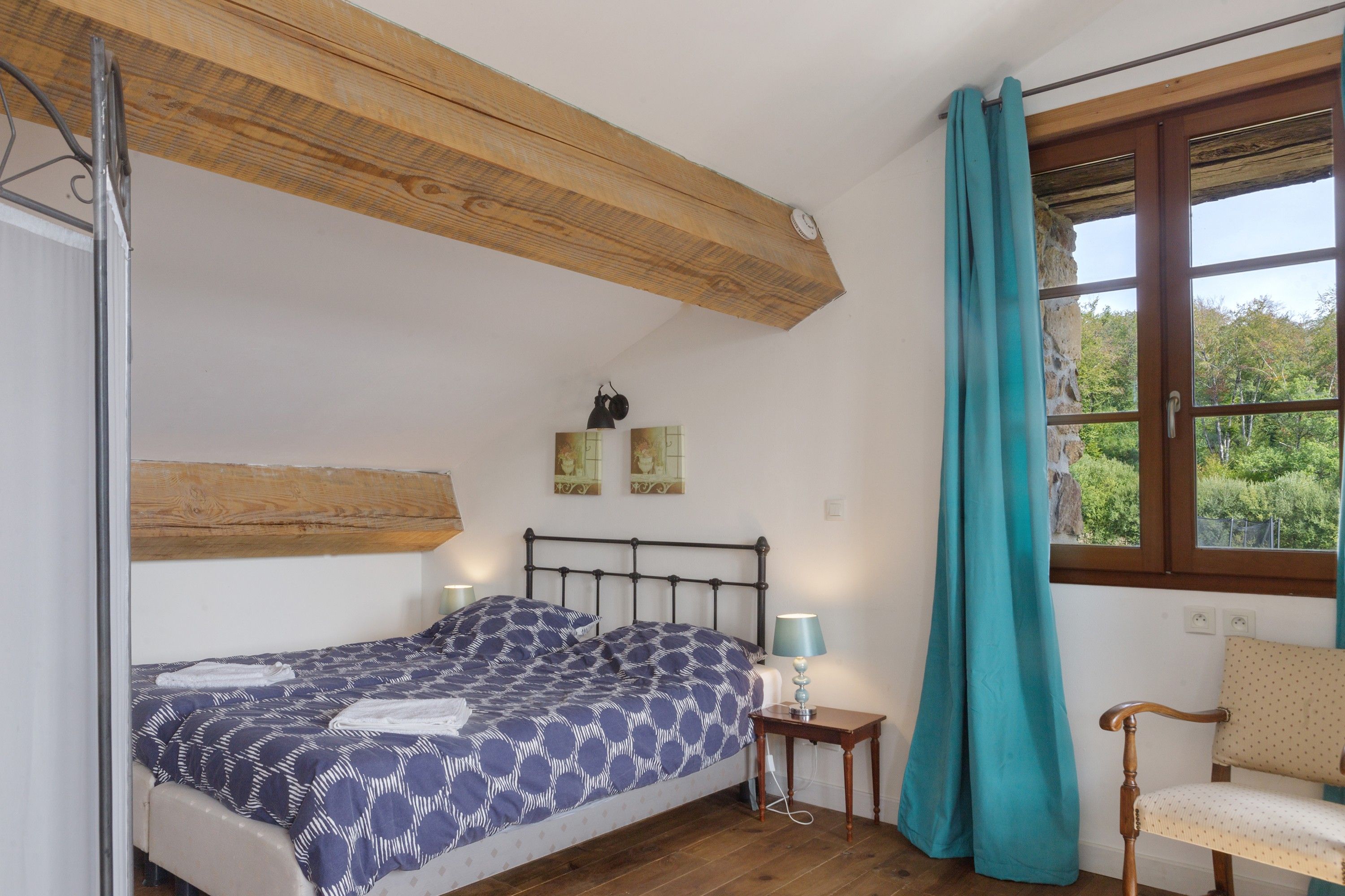 Un dormitorio con cama, mesita de noche, silla, ventana con cortinas y vigas de madera en el techo.