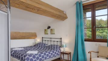 Un dormitorio con cama, mesita de noche, silla, ventana con cortinas y vigas de madera en el techo.