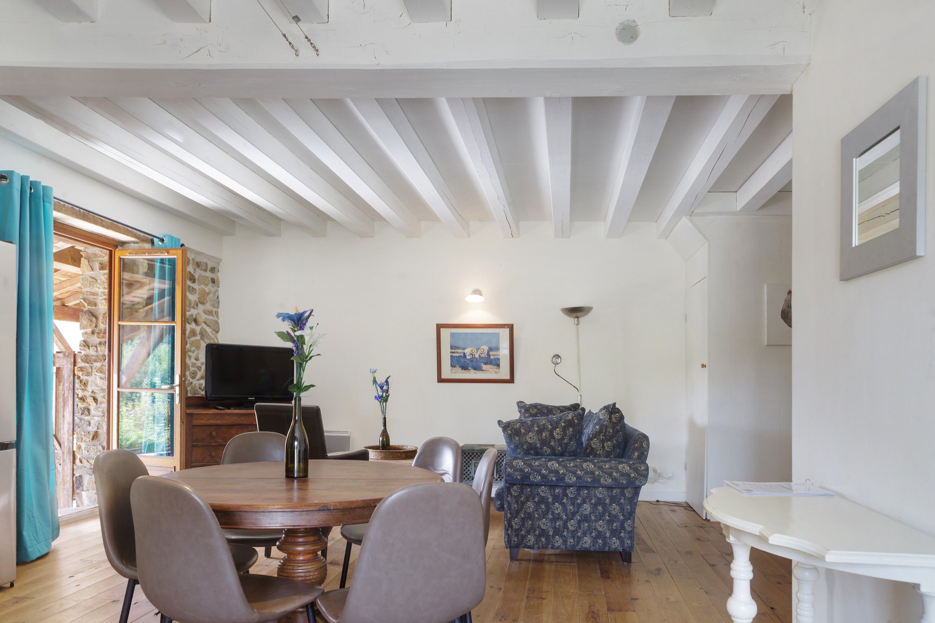 Uma sala de jantar com mesa e cadeiras de madeira, um sofá e uma televisão numa parede de pedra.