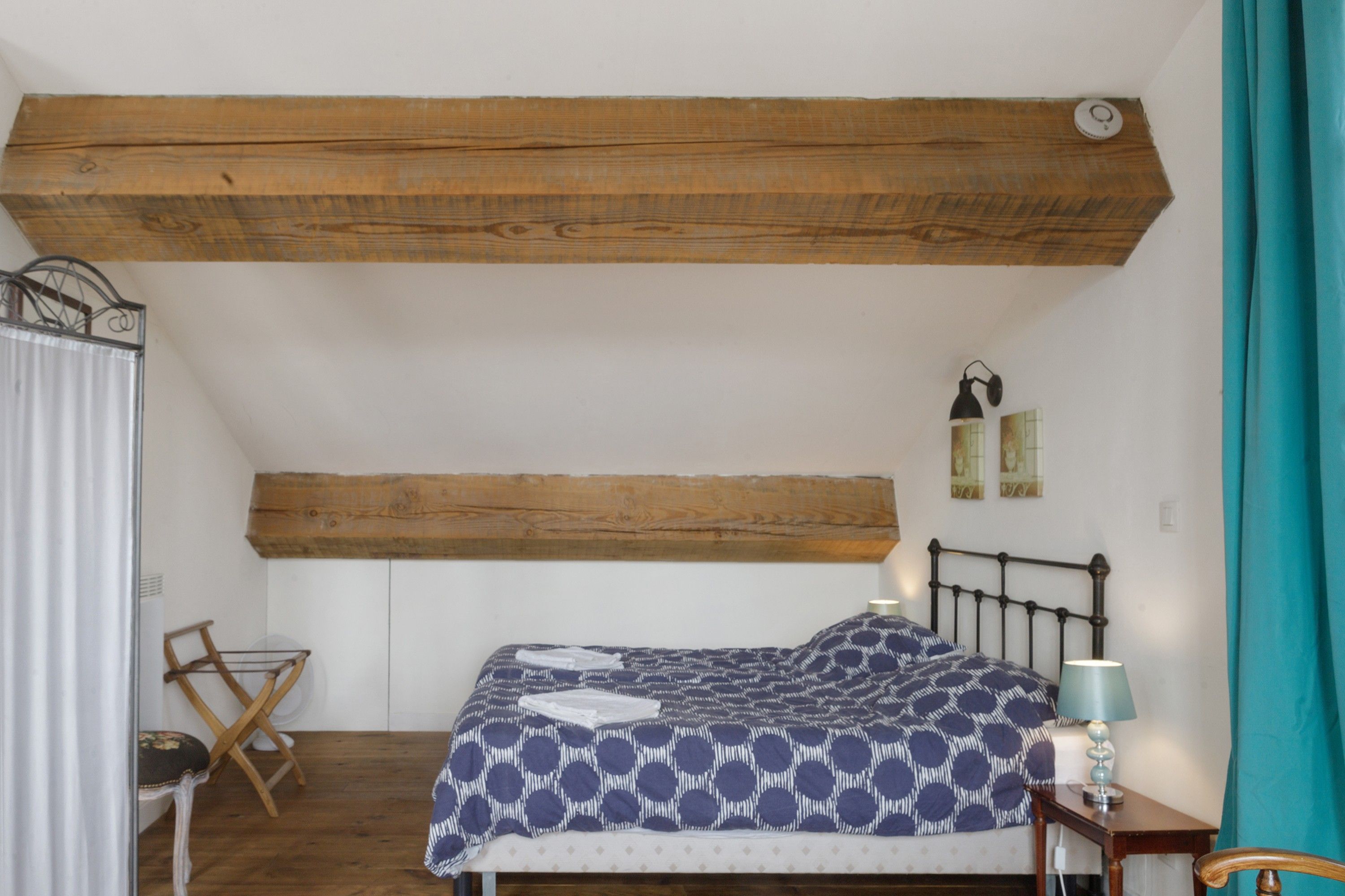 Quarto com teto de madeira, cama com colcha estampada, uma cadeira, mesa pequena com abajur e janela com cortinas.