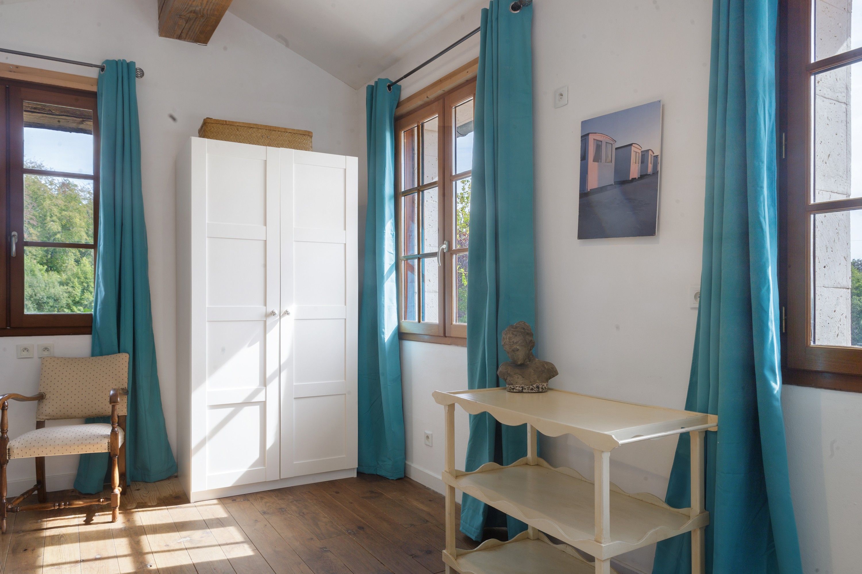 A room with a wooden floor, a white wardrobe, a wooden chair, a small table, and teal curtains.