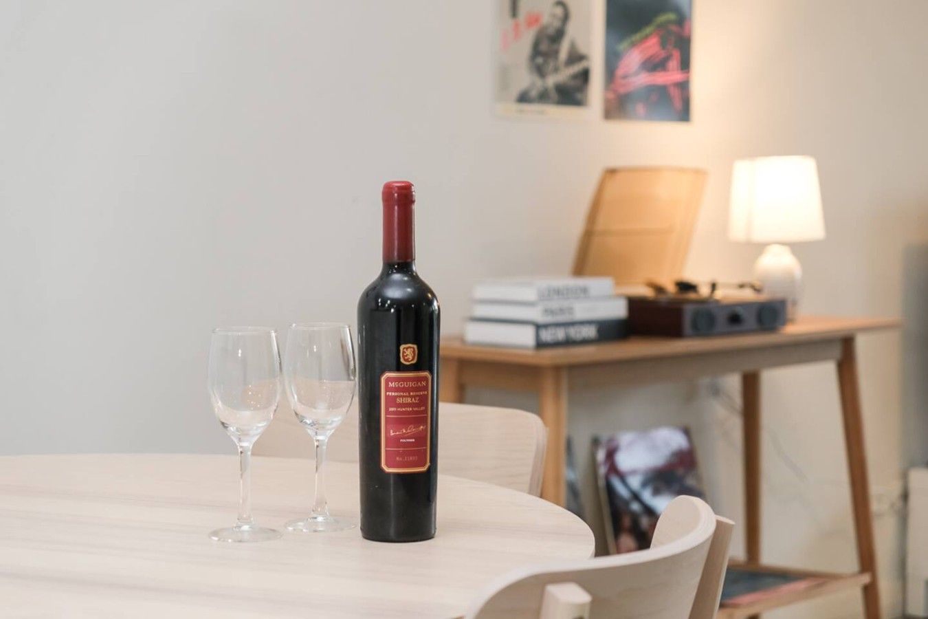 Une bouteille de vin sur une table, avec deux verres, une lampe et des livres en arrière-plan.