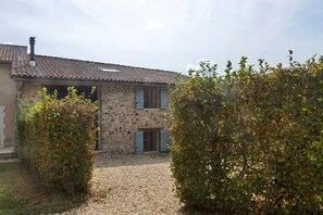A stone house with a tiled roof, a gravel driveway, and a hedge in the foreground.