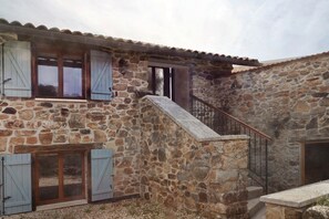 A stone building with a staircase leading to a second floor, featuring wooden shutters and a metal railing.
