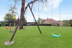 A playground with a swing set and a slide, surrounded by a grassy area and a stone building in the background.