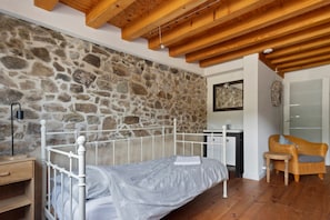 A bedroom with a stone wall, a bed with a white frame, a wooden ceiling, a small desk, a chair, and a door.