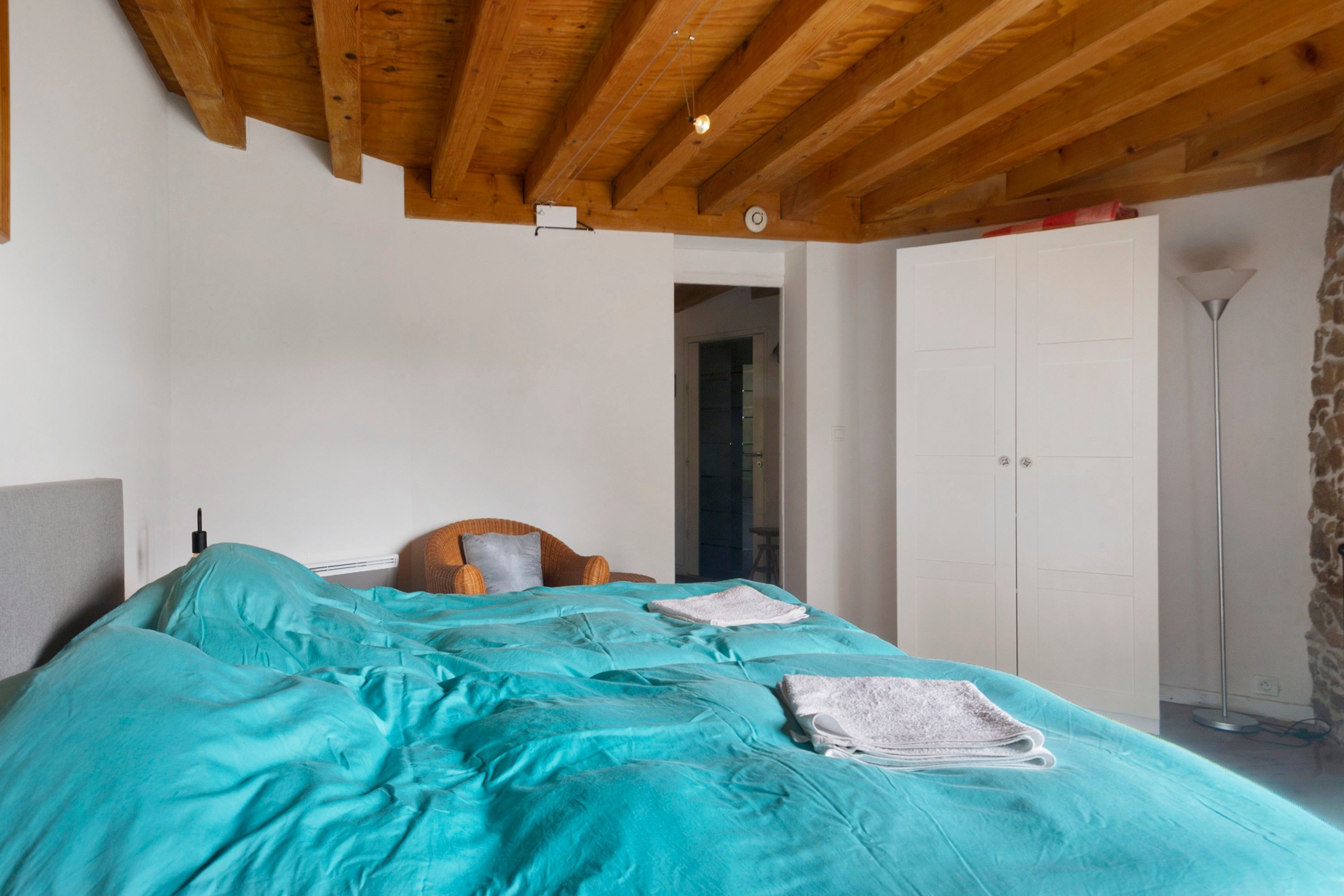 Une chambre avec un plafond en bois, un lit avec une literie de couleur turquoise, une chaise en osier et une armoire.