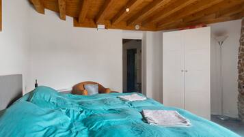 Une chambre avec un plafond en bois, un lit avec une literie de couleur turquoise, une chaise en osier et une armoire.