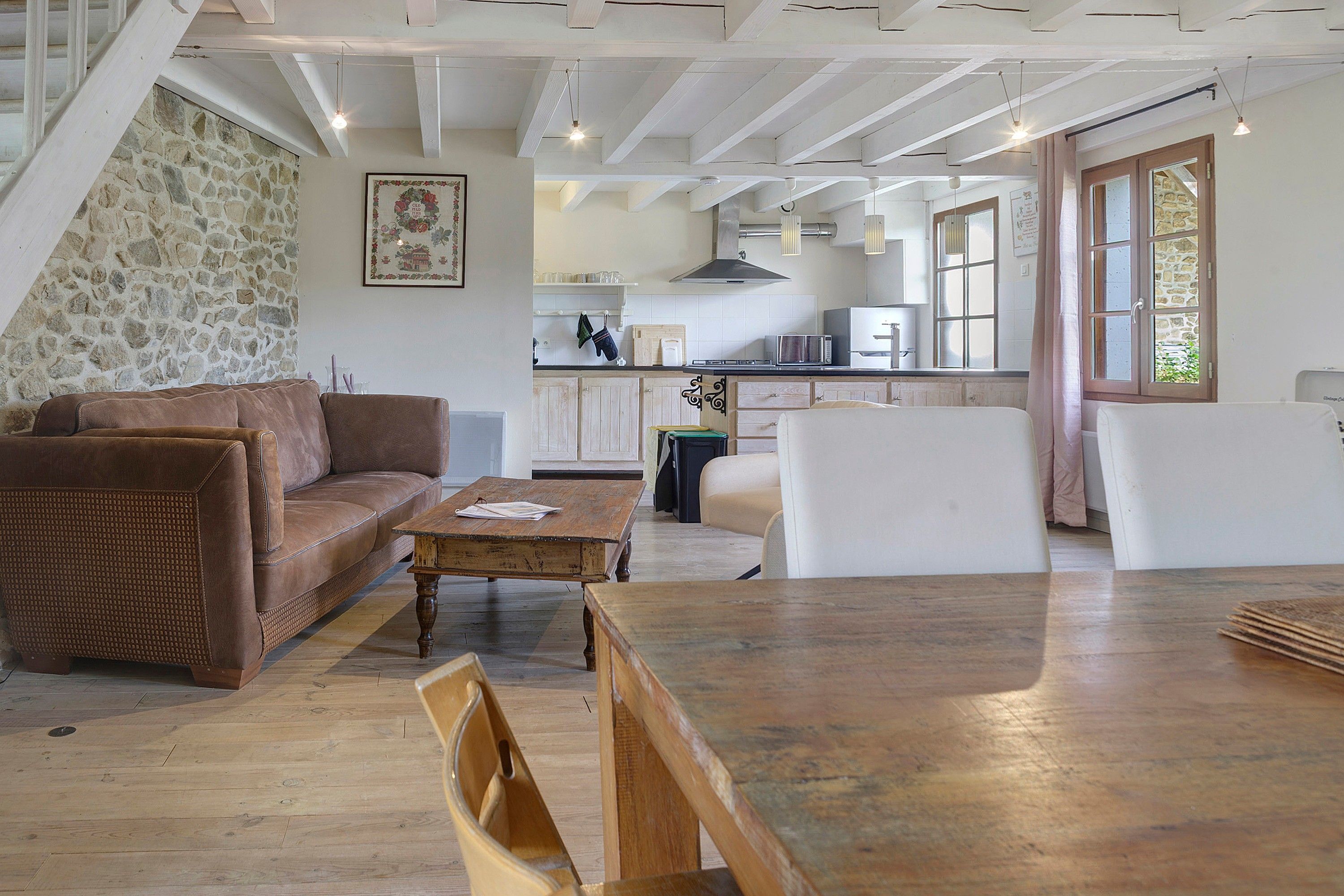 A spacious living area with a brown leather sofa, a wooden coffee table, and a dining table. A kitchen with white cabinets and a stone wall is visible in the background.