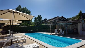 A swimming pool with lounge chairs and umbrellas, surrounded by a garden and a building with a tiled roof.