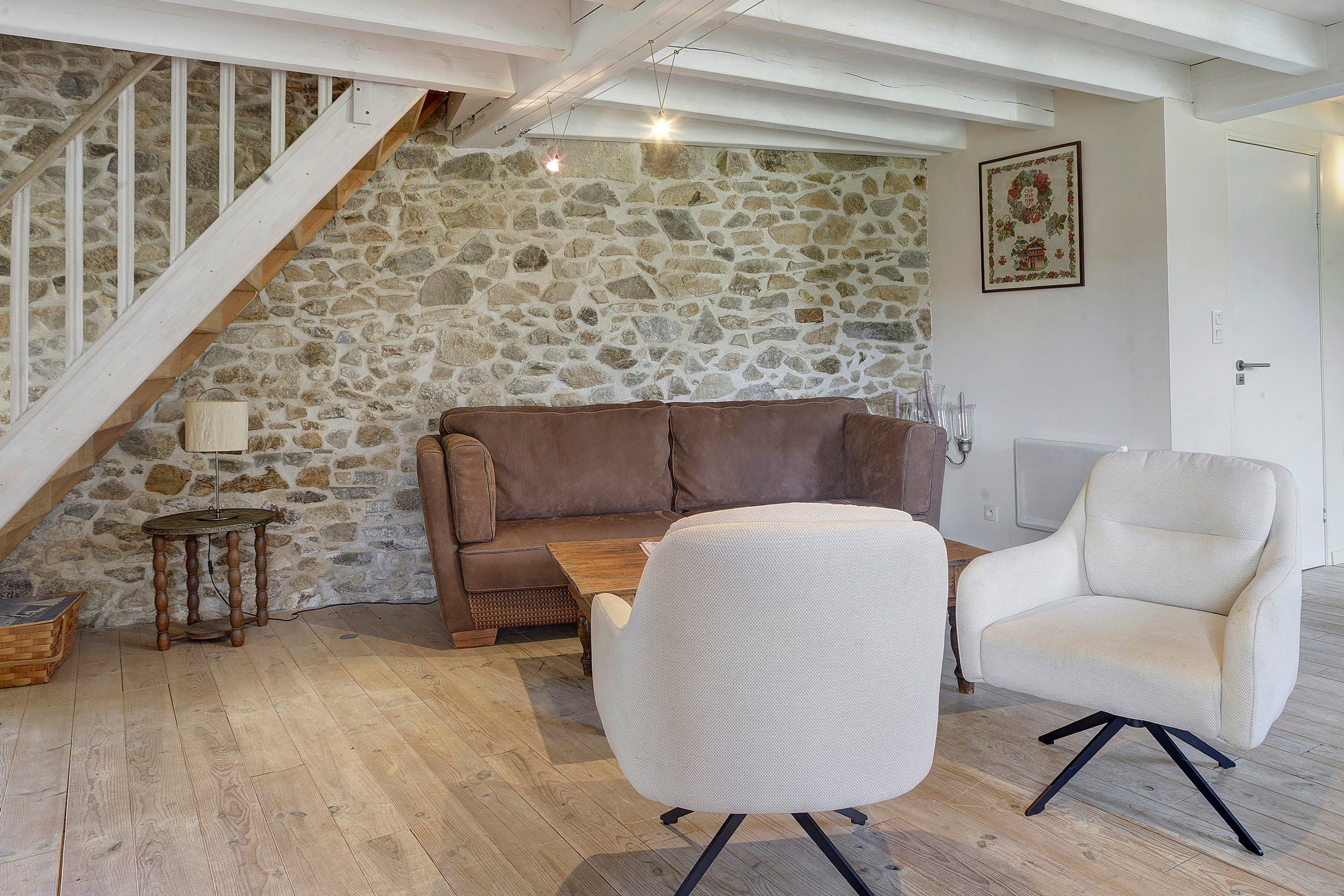 Un salon chaleureux avec un mur en pierre, un canapé en cuir marron, un fauteuil blanc et une table basse en bois.