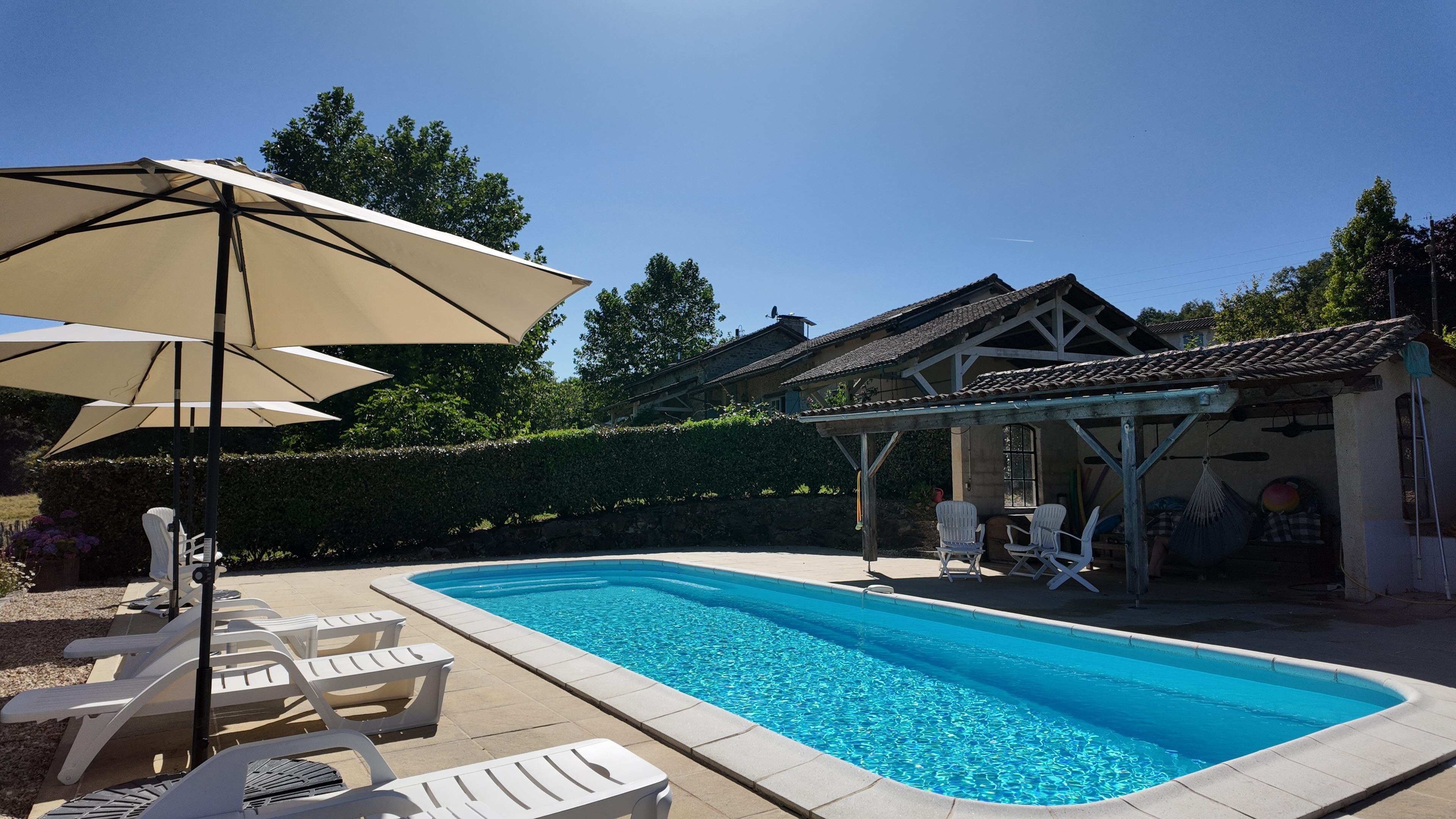 A swimming pool with lounge chairs and umbrellas, surrounded by a garden and a building with a tiled roof.
