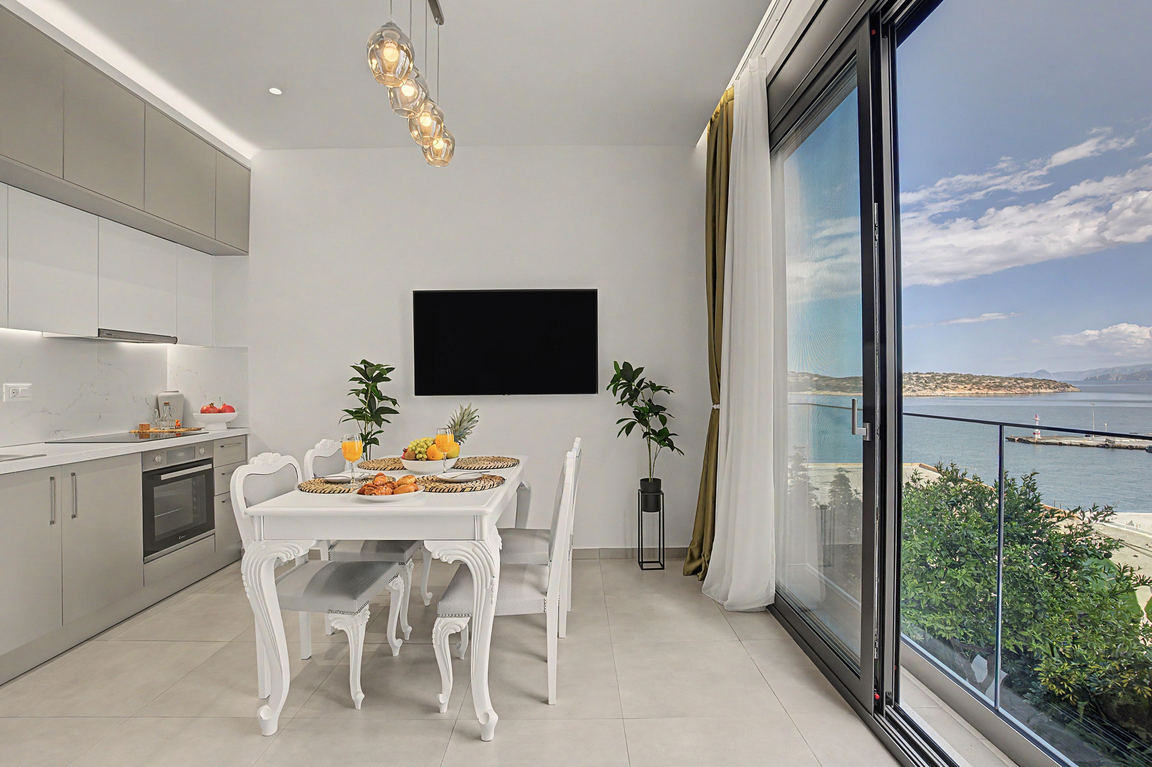 A modern kitchen with white cabinets, a dining table set for two, and a large window offering a view of the sea.