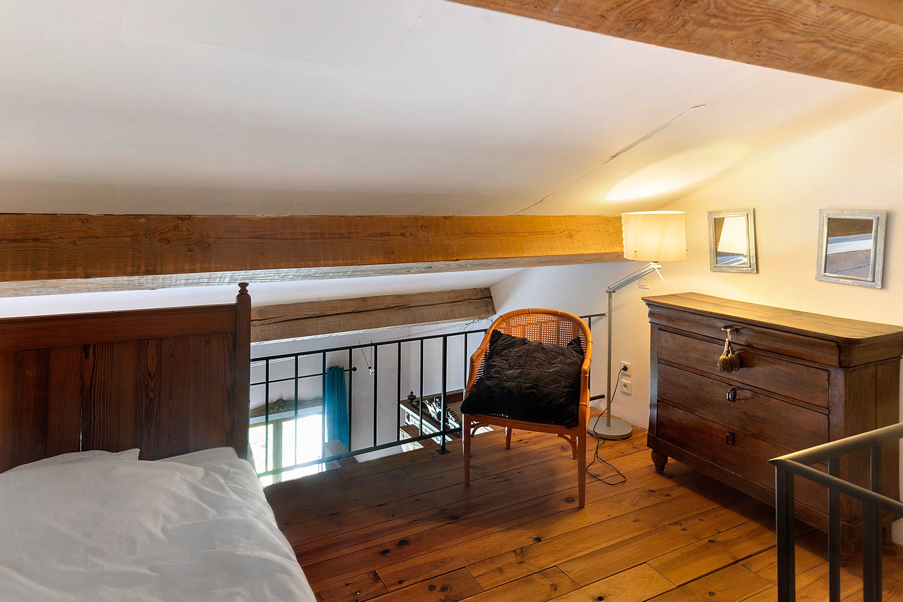 Una habitación con piso de madera, una cama, una silla, un aparador y un balcón con baranda.