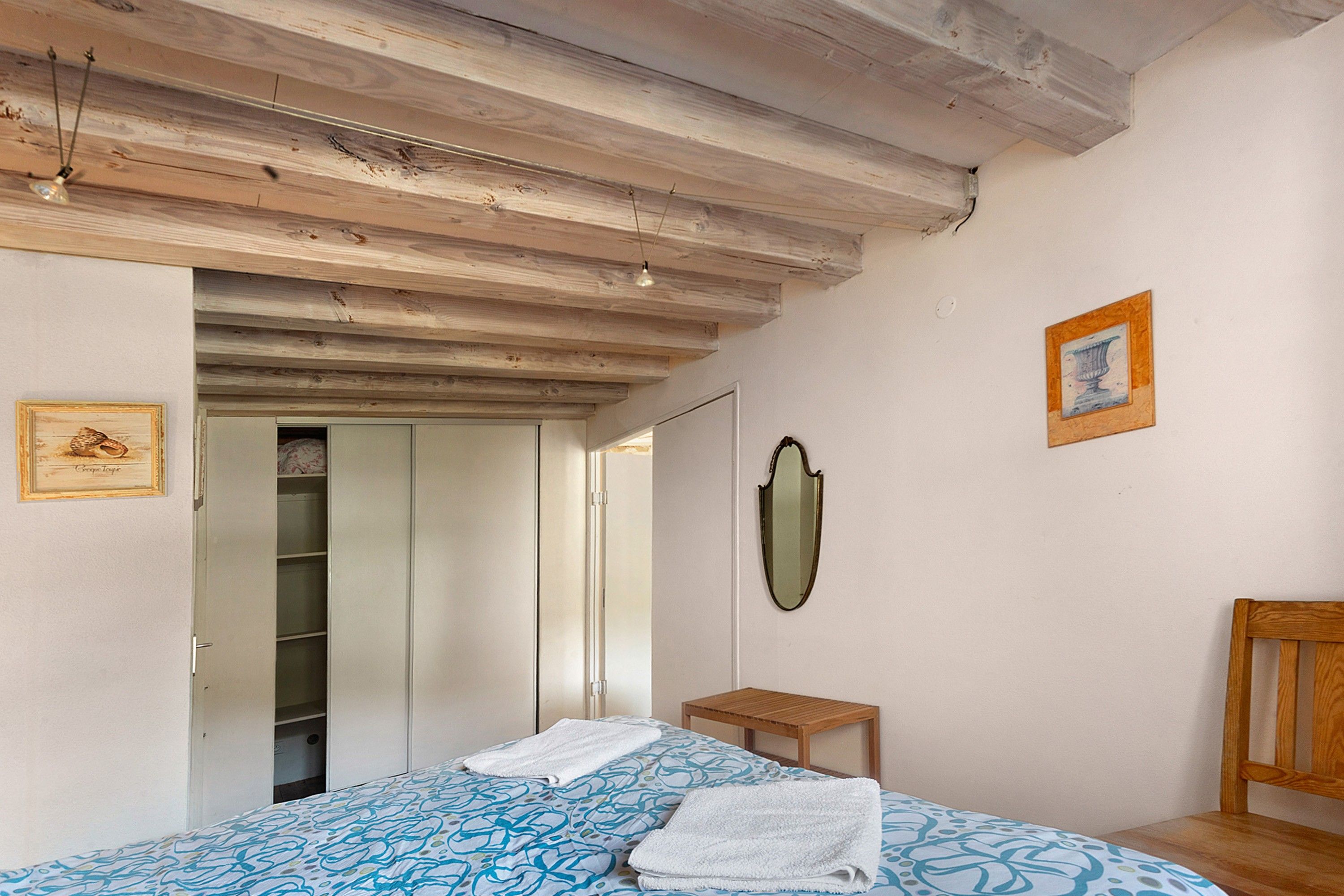 Quarto com cama, cadeira de madeira, mesa de cabeceira, espelho e porta que dá para outro cômodo.