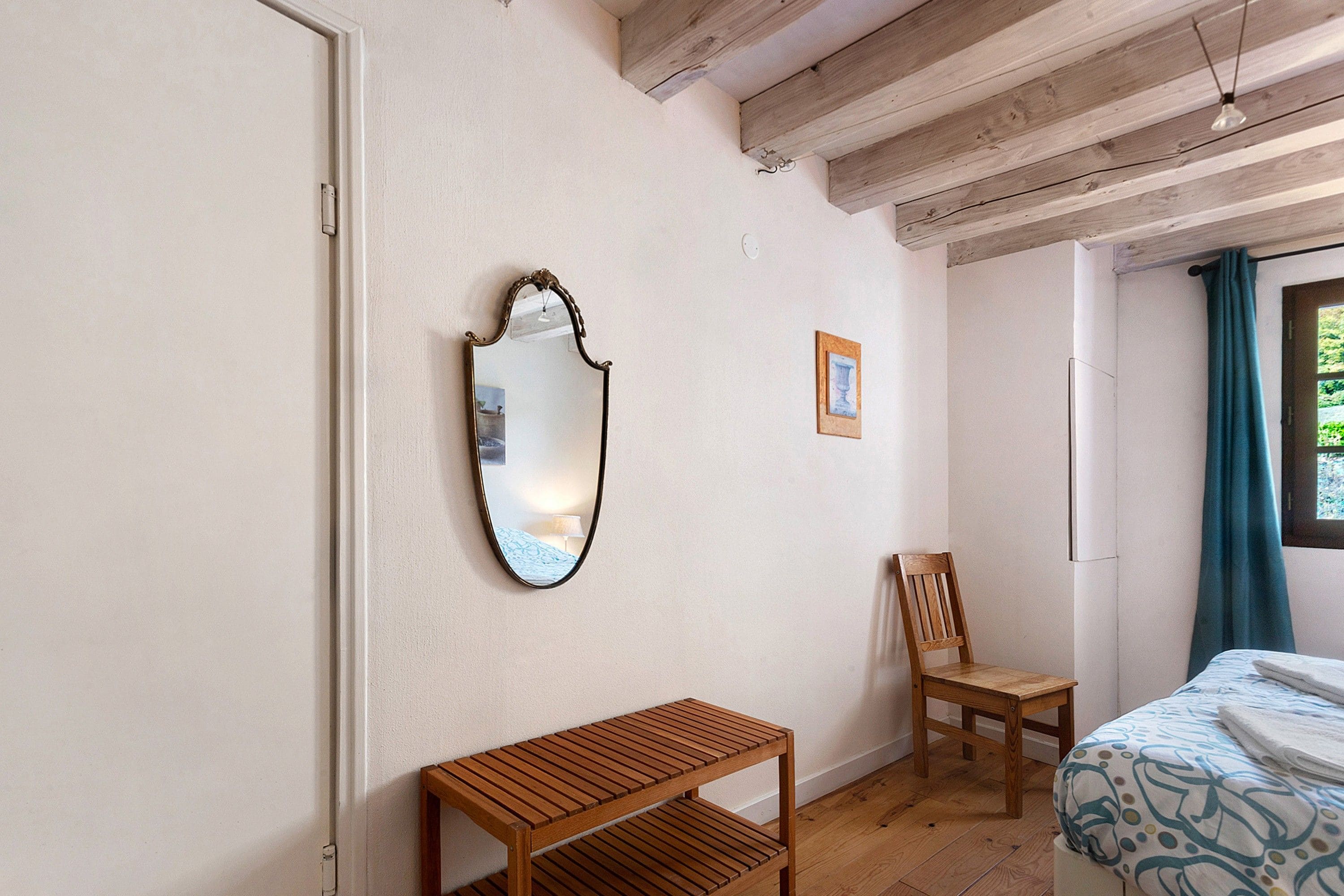 Quarto com um banco de madeira, uma cadeira, uma cama com manta estampada, um espelho e um quadro na parede.