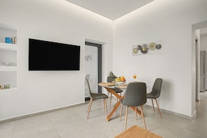 A modern dining area with a flat-screen TV mounted on the wall, a round glass table, and four grey upholstered chairs with wooden legs. - Apartment 'Sea View Uno 1' with Sea View, Wi-Fi and Air Conditioning (Agios Nikolaos)