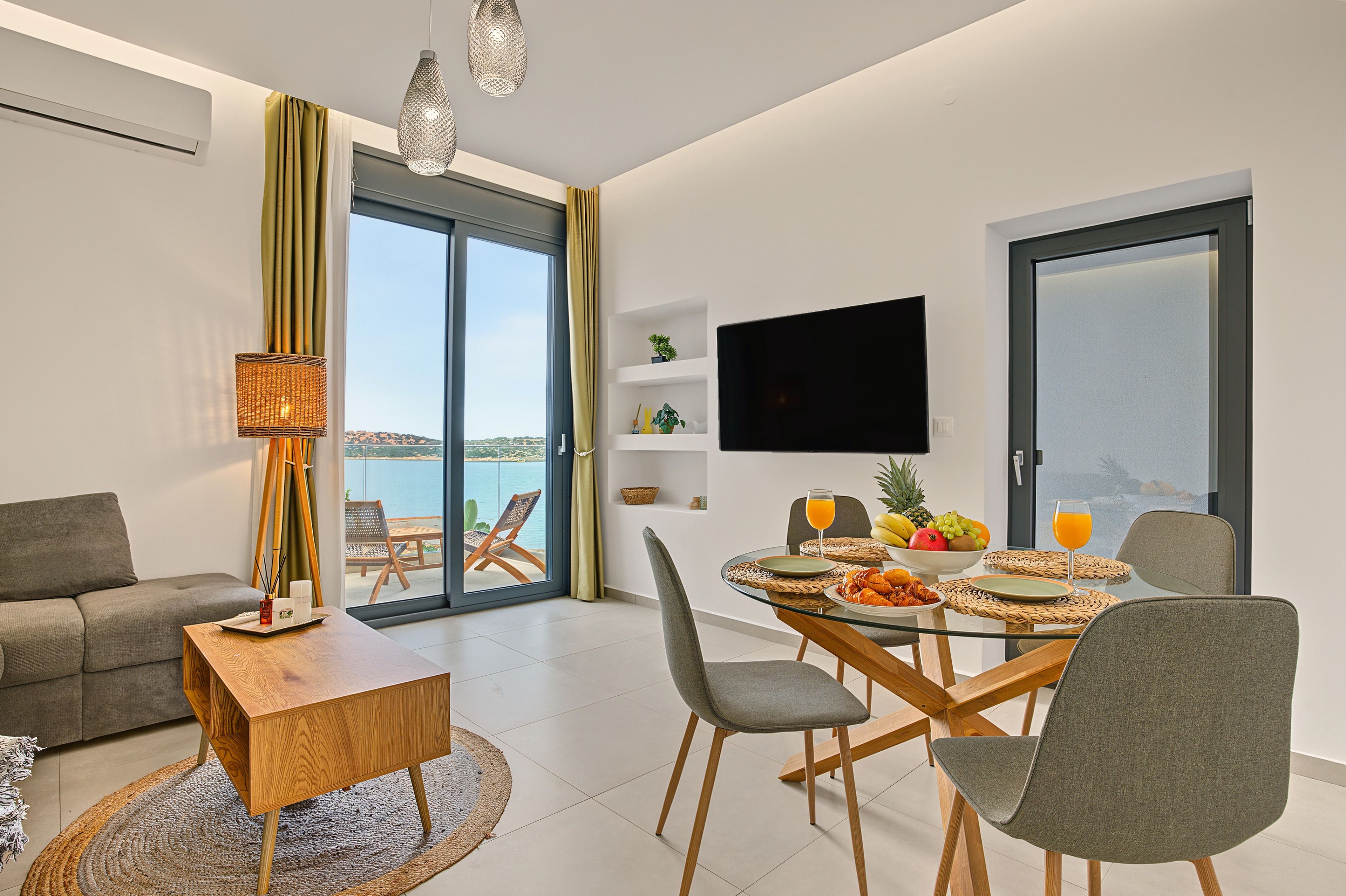 A modern living room with a glass table, grey chairs, a sofa, and a TV mounted on the wall.