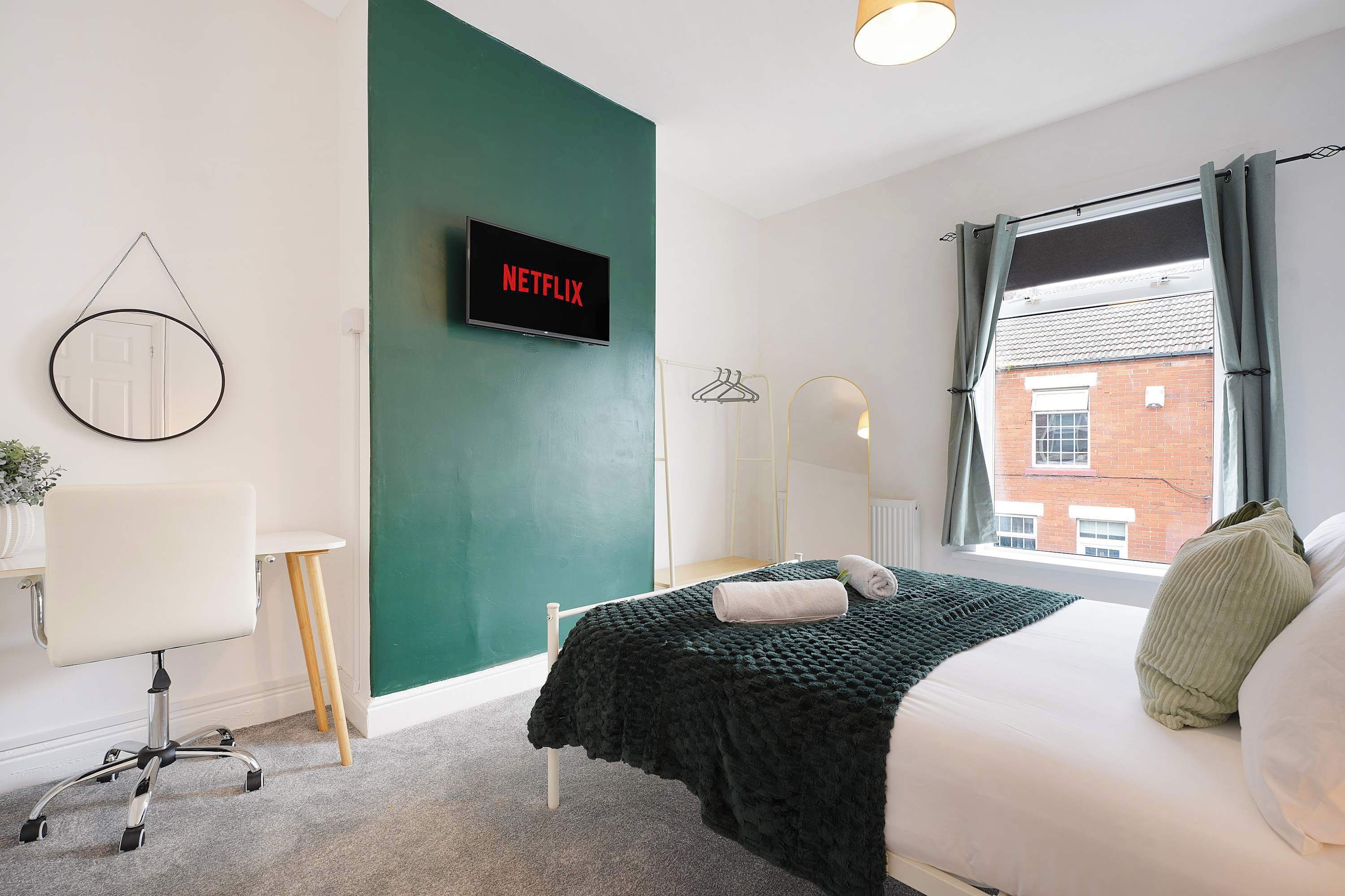 A bedroom with a bed, a desk, a chair, a TV displaying 'NETFLIX,' and a window with curtains.