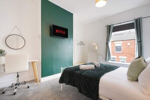 Un dormitori amb un llit, escriptori, cadira, televisió que mostra 'NETFLIX' i una finestra amb cortines.
