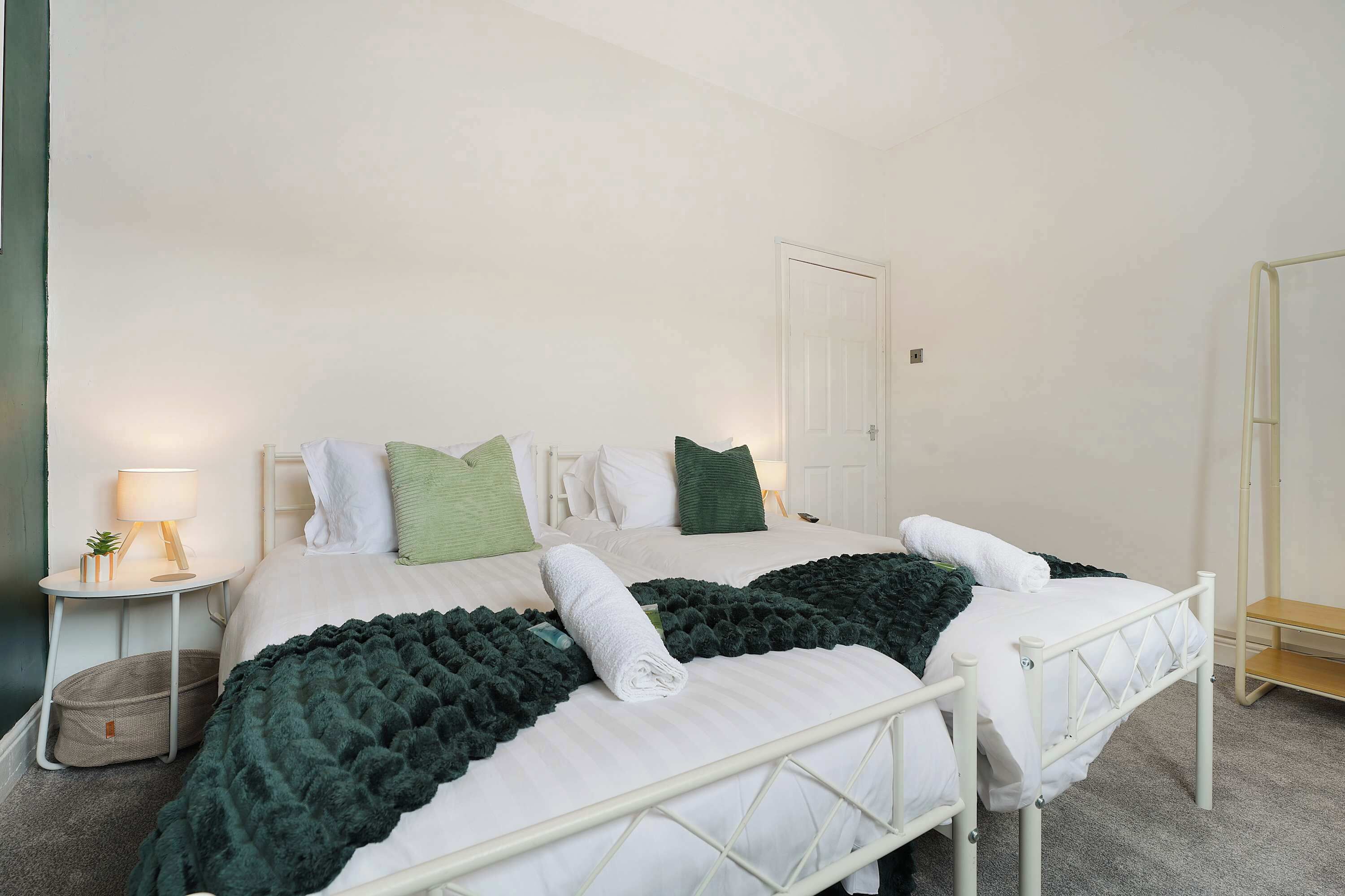 A neatly made bed with white bedding, a green throw blanket, and a white towel. There is a bedside table with a lamp and a small plant. The room has white walls and a door.