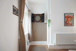 Een kamer met een dartbord, een inlijsting tijdschrift, een radiator en een raam met gordijnen.