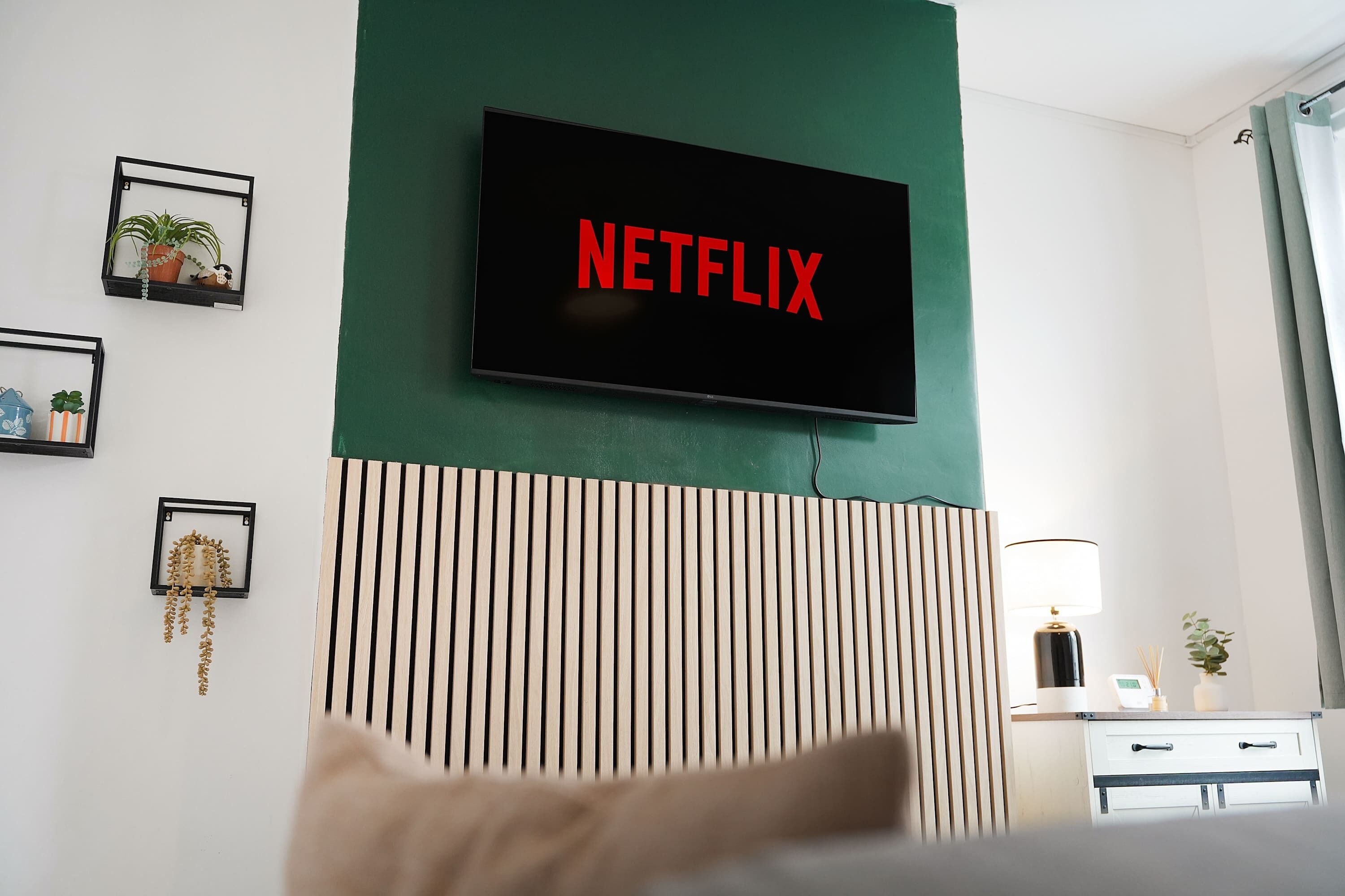 Hotelkamer met een wandtelevisie waarop het Netflix-logo staat, een bed, een nachtkastje met een lamp en een wandplank met decoratieve objecten.
