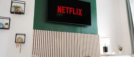 A hotel room with a mounted television displaying the Netflix logo, a bed, a bedside table with a lamp, and a wall-mounted shelf with decorative items.