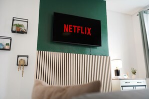Ein Hotelzimmer mit einem an der Wand befestigten Fernseher, der das Netflix-Logo zeigt, einem Bett, einem Nachttisch mit Lampe und einem Wandregal mit Dekoration.