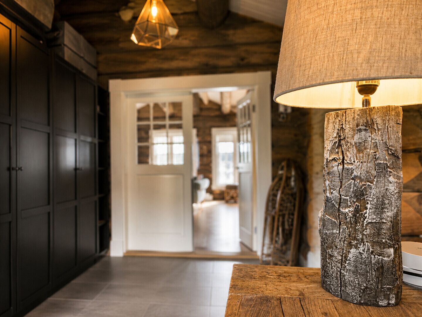 A hallway with a wooden floor, a rustic lamp, and a door leading to a room with a bed.