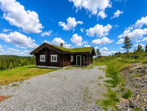 A wooden cabin with a green roof, surrounded by a gravel driveway, set against a backdrop of a clear blue sky and a forested hillside.