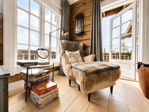 Un cuarto acogedor con pared de madera, una silla confortable, una mesita con libros y una ventana con cortinas.